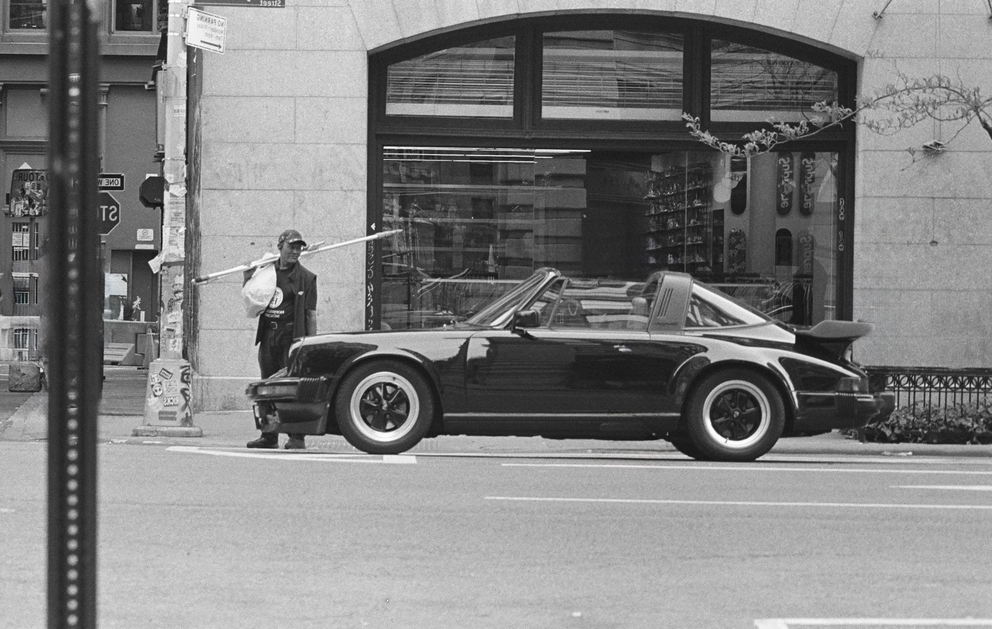 Man in NYC holding window cleaning tools looking at a black Porsche. The pedestrians and the cone in front of the car have been edited out. It blew my mind when i first did this that cleaning up the cone also removed the reflection of the cone on the exterior.